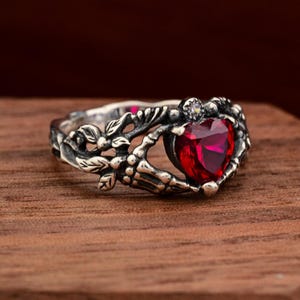 May include: A silver ring with a heart-shaped, deep red gemstone. The ring features a detailed, ornate design with floral and skeletal elements. A small, clear stone accents the top of the heart. The ring is set against a wooden background.