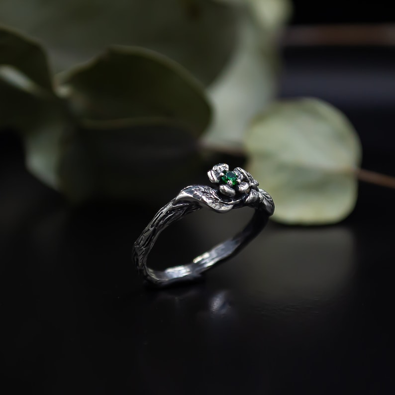 May include: A silver ring with a green gemstone set in a flower design. The ring is shaped like a twisted branch.