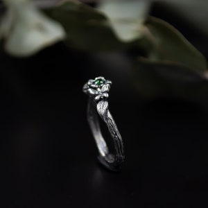 May include: A silver ring with a green gemstone and a twig-like band. The ring is designed to look like a branch with leaves and a flower.