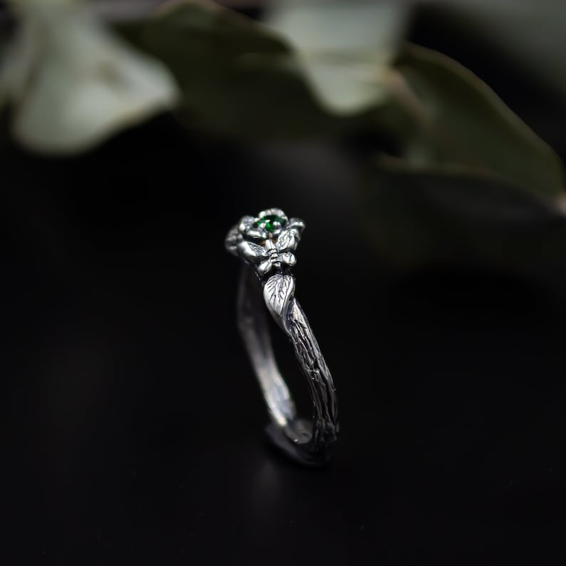 May include: A silver ring with a green gemstone and a twig-like band. The ring is designed to look like a branch with leaves and a flower.