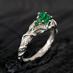 May include: A silver ring with a leafy design and three green gemstones. The ring is set on a dark background.