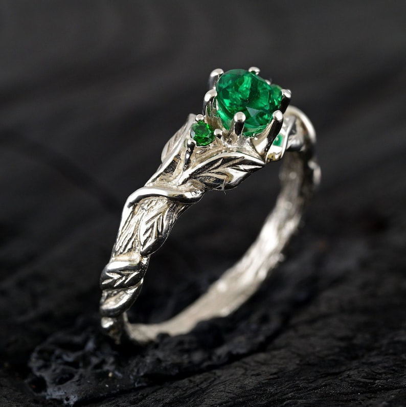 May include: A silver ring with a leafy design and three green gemstones. The ring is set on a dark background.