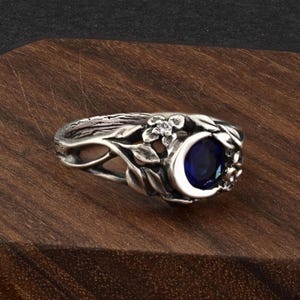 May include: A silver ring featuring a crescent moon design with a deep blue gemstone. The ring is adorned with floral and leaf details, creating an elegant and nature-inspired aesthetic. The ring is set on a wooden surface.