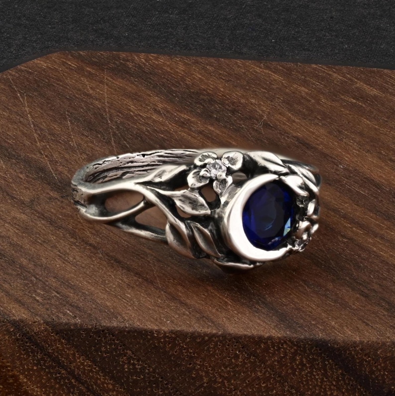 May include: A silver ring featuring a crescent moon design with a deep blue gemstone. The ring is adorned with floral and leaf details, creating an elegant and nature-inspired aesthetic. The ring is set on a wooden surface.