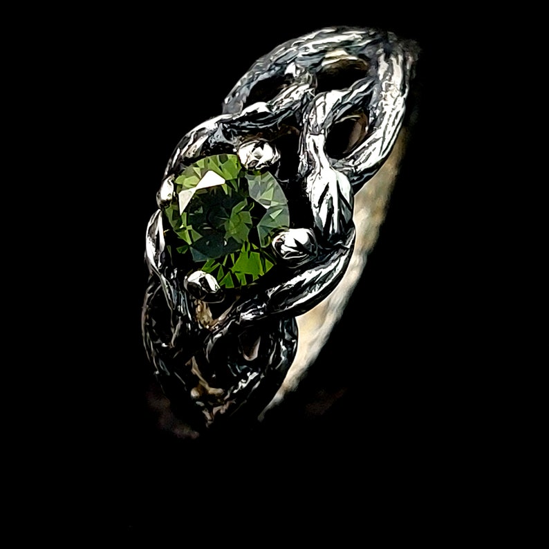 May include: A silver ring with a green gemstone set in a twisted, organic design. The ring is reminiscent of a tree branch or vine.