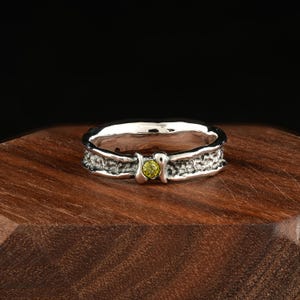 May include: A silver ring with a textured band and a small, round, light green gemstone. The ring is set on a dark wooden surface, with a black background.