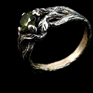 May include: A silver ring with a green gemstone set in a branch-like design. The ring is made to look like a tree branch with leaves.