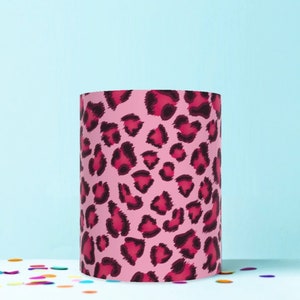 May include: A cylindrical lampshade featuring a pink leopard print design with dark pink and black spots. The lampshade is set against a light blue background, with colorful confetti scattered at the base.