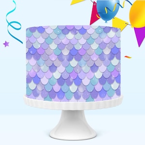May include: A round cake with a mermaid scale design in shades of purple, blue, and white sits on a white cake stand. The cake is decorated with a repeating pattern of overlapping scales. Party decorations, including balloons and streamers, are in the background.