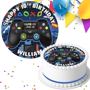 May include: A round cake and matching plate with a video game controller design. The cake and plate feature the text "Happy 10th Birthday William" and "Game On Level Up". The cake has white icing and a blue ribbon.