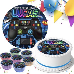 Op de afbeelding: Een ronde taarttopper en bijpassende cupcaketoppers met een videogame-thema, een zwarte controller, de tekst HAPPY 10TH BIRTHDAY LUCAS en GAME ON LEVEL UP. Het ontwerp bevat blauwe, gele en rode accenten op een donkere achtergrond.