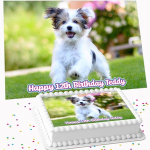 May include: A white birthday cake decorated with an image of a puppy running on grass. The cake has a white, piped border and the text "Happy 12th Birthday Teddy". Confetti in various colours is scattered around the cake.