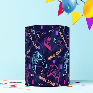 May include: A cylindrical party decoration with a dark blue background and a repeating pattern of video game controllers, game consoles, and the words "LET'S PLAY" and "GAME ZONE". The decoration is surrounded by party decorations.