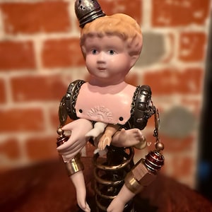 Könnte beinhalten: Eine einzigartige Steampunk-Puppenskulptur mit einem Porzellanpuppenkopf mit Metallhelm. Der Puppenkörper besteht aus Metallfedern, Zahnrädern und Hardware, mit kleinen Puppenhänden. Die Skulptur hat eine Vintage- und Industrieästhetik.