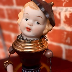May include: A vintage porcelain doll head with a brown hat and a gold-toned collar. The doll has blonde hair, red lips, and is attached to a decorative body with gold and brown accents. The doll's arms are adorned with gold bracelets and dangling beads.