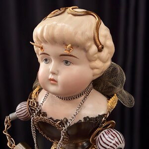 Puede incluir: Busto de muñeca de porcelana con elaborados adornos de estilo steampunk. La muñeca tiene cabello rubio adornado con espirales de metal dorado y pequeños cuernos. Un collar de cadena plateada y elementos decorativos están unidos al busto.