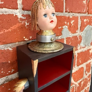 Könnte beinhalten: Einzigartiges, handgefertigtes Regal mit einem Vintage-Puppenkopf mit blauen Augen und goldfarbenem Haar. Das Regal hat einen dunklen Rahmen mit roten Einlegeböden, goldfarbenen Eckdetails und einem Puppenarm. Das Regal ist an einer Backsteinmauer befestigt. Höhe ca. 60 cm.