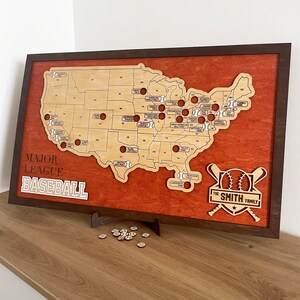 May include: A wooden map of the United States with baseballs marking the location of Major League Baseball stadiums. The map is framed in brown wood and has a red background. The text "MAJOR LEAGUE BASEBALL" is printed in black at the bottom left corner. The text "THE SMITH FAMILY" is printed in black at the bottom right corner.
