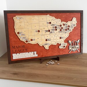 May include: A wooden map of the United States with baseballs marking the locations of Major League Baseball teams. The map is framed in a dark wood frame and has a red background. The text "MAJOR LEAGUE BASEBALL" is printed below the map. The text "THE SMITH FAMILY" is printed below a baseball and bats.
