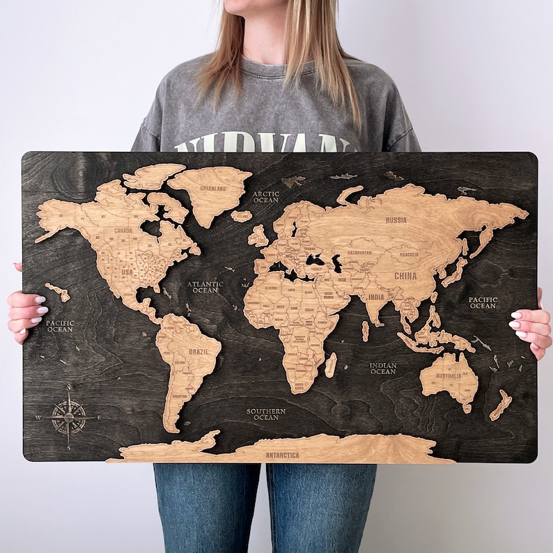 May include: A wooden world map with a black background and brown continents. The map is detailed with country borders and names of oceans. The map is being held by a person's hands.