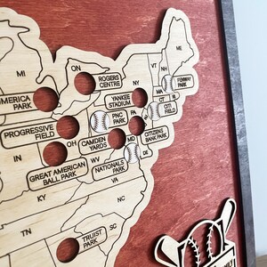May include: A wooden map of the United States with baseball stadiums marked. Each stadium is represented by a small circle with a baseball inside. The map is stained a dark reddish brown color. The map is surrounded by a dark brown frame.
