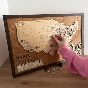 May include: A wooden map of the United States with baseball stadiums marked. The map is framed in brown wood and has the text "Major League Baseball" printed below the map. A hand is placing a baseball-shaped marker on the map.