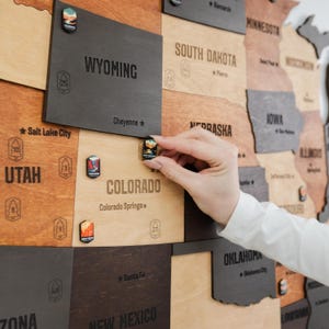 May include: A wooden wall map of the United States featuring laser-engraved state names and capital cities. Small, colorful hexagonal pins depicting national parks are placed on the map, with one pin being positioned on Colorado.