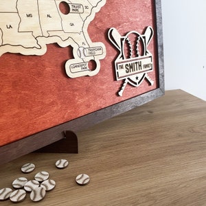 May include: A wooden baseball map with a red stain finish. The map is framed in dark wood and has a stand. The map features baseball stadiums across the United States. The map also includes a baseball-themed logo with the text "THE SMITH FAMILY".