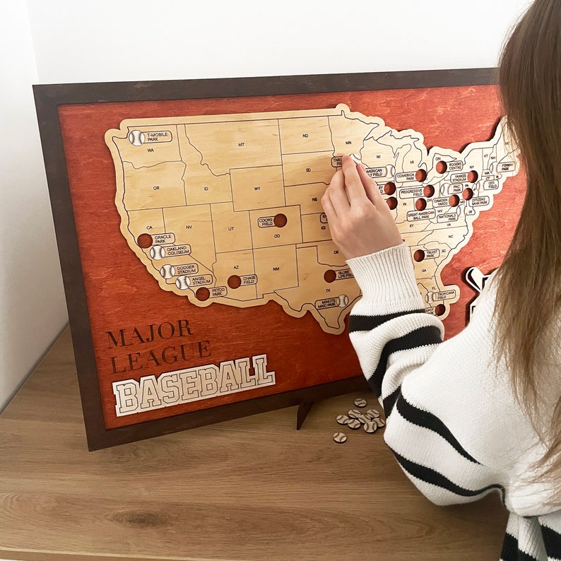 May include: A wooden map of the United States with a red background and a light wood outline of the states. The map is labeled "Major League Baseball" and has small baseballs marking the location of each major league baseball stadium. A person's hand is placing a small white baseball on the map.
