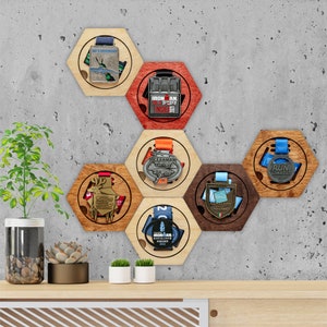 May include: A hexagonal wooden medal display with six different medals. The medals are from various races, including IRONMAN, OCEANMAN, and RUN LIMASSOL. The display is mounted on a gray wall.