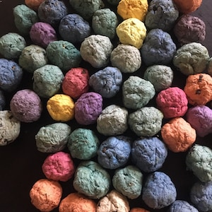 May include: A selection of multi-coloured seed bombs, with shades of orange, blue, yellow, and green. The seed bombs have a textured, organic look, implying they are handmade. They are displayed on a dark surface.