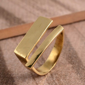 Könnte beinhalten: Ein goldfarbener Ring mit modernem, offenem Design. Der Ring hat einen rechteckigen Balken, der sich über den Finger erstreckt und mit einem Band verbunden ist. Die polierte Oberfläche des Rings reflektiert das Licht und hebt seine geometrische Form hervor.