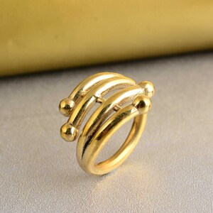 Könnte beinhalten: Ein goldener Ring mit einem einzigartigen Design, das drei geschwungene Bänder mit abgerundeten Enden aufweist. Die Bänder sind miteinander verschlungen und erzeugen einen modernen und geometrischen Look.