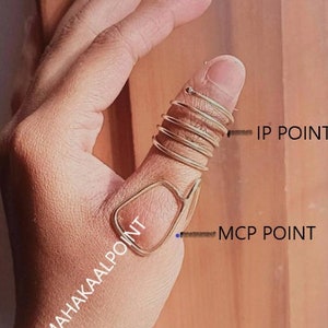 Könnte beinhalten: Nahaufnahme einer Hand, die einen goldenen Ring am Daumen trägt. Der Ring besteht aus mehreren Metallbändern, die zusammengerollt sind. Das Bild zeigt die Position des IP-Punkts und des MCP-Punkts am Daumen.
