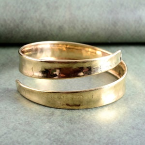 Könnte beinhalten: Ein goldfarbenes Metall-Armreif mit gehämmerter Oberfläche. Das Armband besteht aus zwei Metallteilen, die umeinander gewickelt sind.