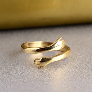 Könnte beinhalten: Ein goldener Ring mit zwei Händen, die sich umeinander schlingen. Der Ring ist aus Metall und hat eine glatte Oberfläche.