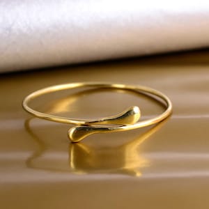 Könnte beinhalten: Ein goldfarbenes Armreif-Armband mit einem einzigartigen, geschwungenen Design. Das Armband ist aus dünnem, glattem Metall gefertigt und hat einen schlichten, eleganten Look.