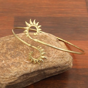 Könnte beinhalten: Ein goldfarbenes Armband aus Metall mit offenem Design und zwei spiralförmigen Enden. Jede Spirale ist mit strahlenförmigen, spitzen Zacken verziert, die an eine Sonne erinnern. Das Armband hat eine glatte, polierte Oberfläche und eine flexible Form.