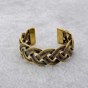 Könnte beinhalten: Ein goldfarbenes Metallarmband mit geflochtenem Design. Das Armband ist offen und hat einen schlichten, eleganten Stil.