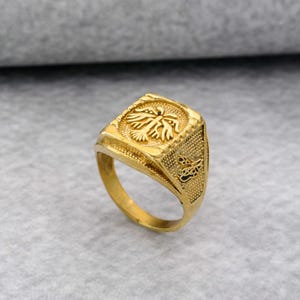 Könnte beinhalten: Ein goldfarbener Ring mit quadratischer Fassung, verziert mit einem geprägten Vogel- oder Adlerdesign. Der Ring hat ein strukturiertes Band und dekorative Seitenelemente. Der Ring ist wahrscheinlich aus Metall und wird vor neutralem Hintergrund gezeigt.