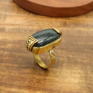 Könnte beinhalten: Ein goldfarbener Ring mit einem großen, rechteckigen, dunkelgrauen Stein, der horizontal eingesetzt ist. Die Metallschiene weist an den Seiten und am Ringkörper komplexe, geprägte Blumenmuster auf, die dem Schmuckstück eine strukturierte Optik verleihen.
