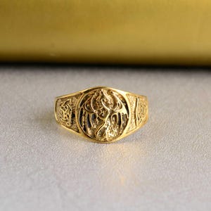 Könnte beinhalten: Ein goldener Ring mit einem detaillierten Drachenmotiv. Der Ring ist ein schlichtes Band mit einem erhabenen runden Design, das einen Drachen zeigt.