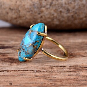 Puede incluir: Un anillo de oro con una gran piedra turquesa de forma ovalada. La piedra preciosa presenta un patr&oacute;n de m&aacute;rmol azul y dorado.
