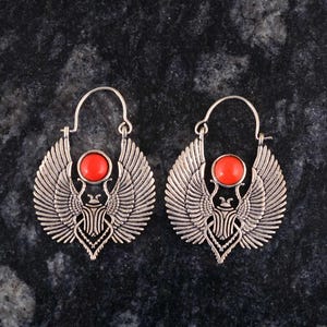 Könnte beinhalten: Ein Paar silberfarbene Ohrringe mit einem detaillierten Flügeldesign und einem zentralen roten Stein. Die Ohrringe weisen ein stilisiertes Skarabäus-Motiv mit aufwendigen Federdetails auf den Flügeln auf.