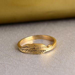 Könnte beinhalten: Ein goldfarbener Ring mit einem Federdesign. Die Feder ist detailliert und hat eine strukturierte Oberfläche.