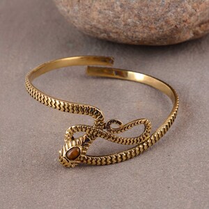 Könnte beinhalten: Ein goldfarbenes Schlangenarmband mit einem braunen Edelsteinauge. Das Armband ist aus Metall und hat ein strukturiertes Design.