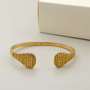 Könnte beinhalten: Ein goldfarbener Metall-Armreif mit offenem Design und zwei abgeflachten, abgerundeten Enden. Die Oberfläche des Armreifs ist mit eingravierten geometrischen Mustern verziert, darunter vertikale Linien, Zickzackmuster und kleine kreisförmige Motive.
