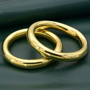 Könnte beinhalten: Zwei polierte goldfarbene Metallringe mit einer glatten, abgerundeten Oberfläche. Die Ringe sind kreisförmig und weisen ein glänzendes, reflektierendes Finish auf, das das Licht einfängt und ein helles, gleichmäßiges Erscheinungsbild erzeugt.