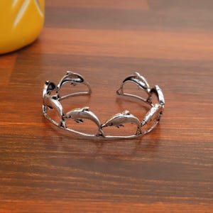 May include: Silver-toned cuff bracelet featuring a repeating dolphin design. The open-ended bracelet is made of metal and has a polished finish. The dolphins are arranged in a circular pattern around the wrist.