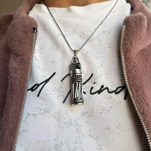 Può includere: Una collana con catena d'argento con un ciondolo d'argento a forma di piccola bottiglia decorata. La bottiglia ha un design dettagliato ed è chiusa con un tappo.